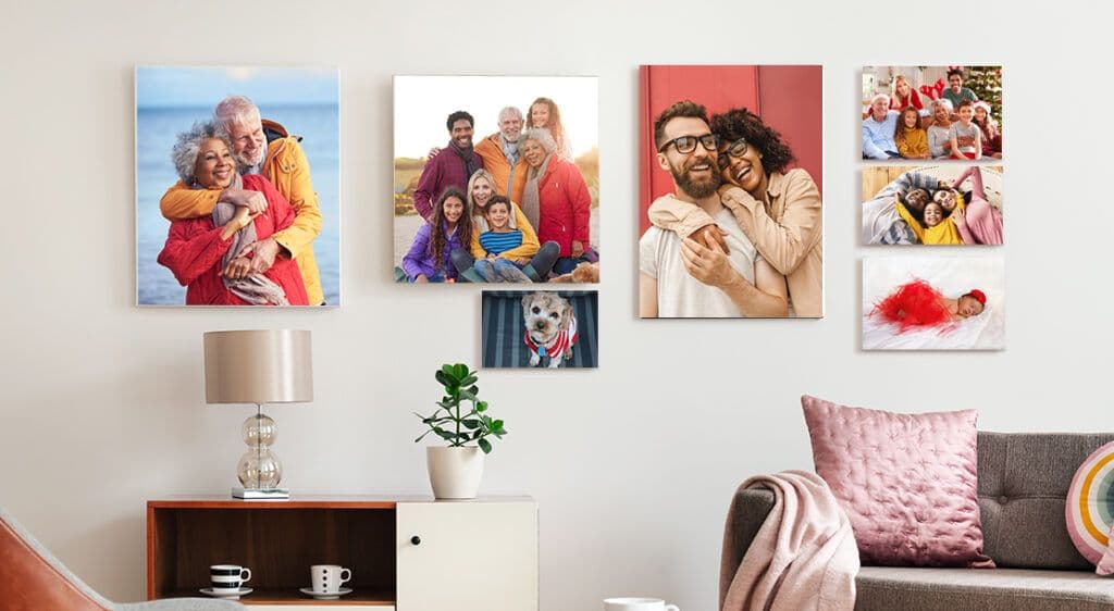 A living room wall displays a collection of family photos printed on canvas in varying sizes. Featured on a white wall, the canvas prints showcase a portrait of grandparents, a young couple, a large family group photo, a brand new infant, and a photo of the family dog.