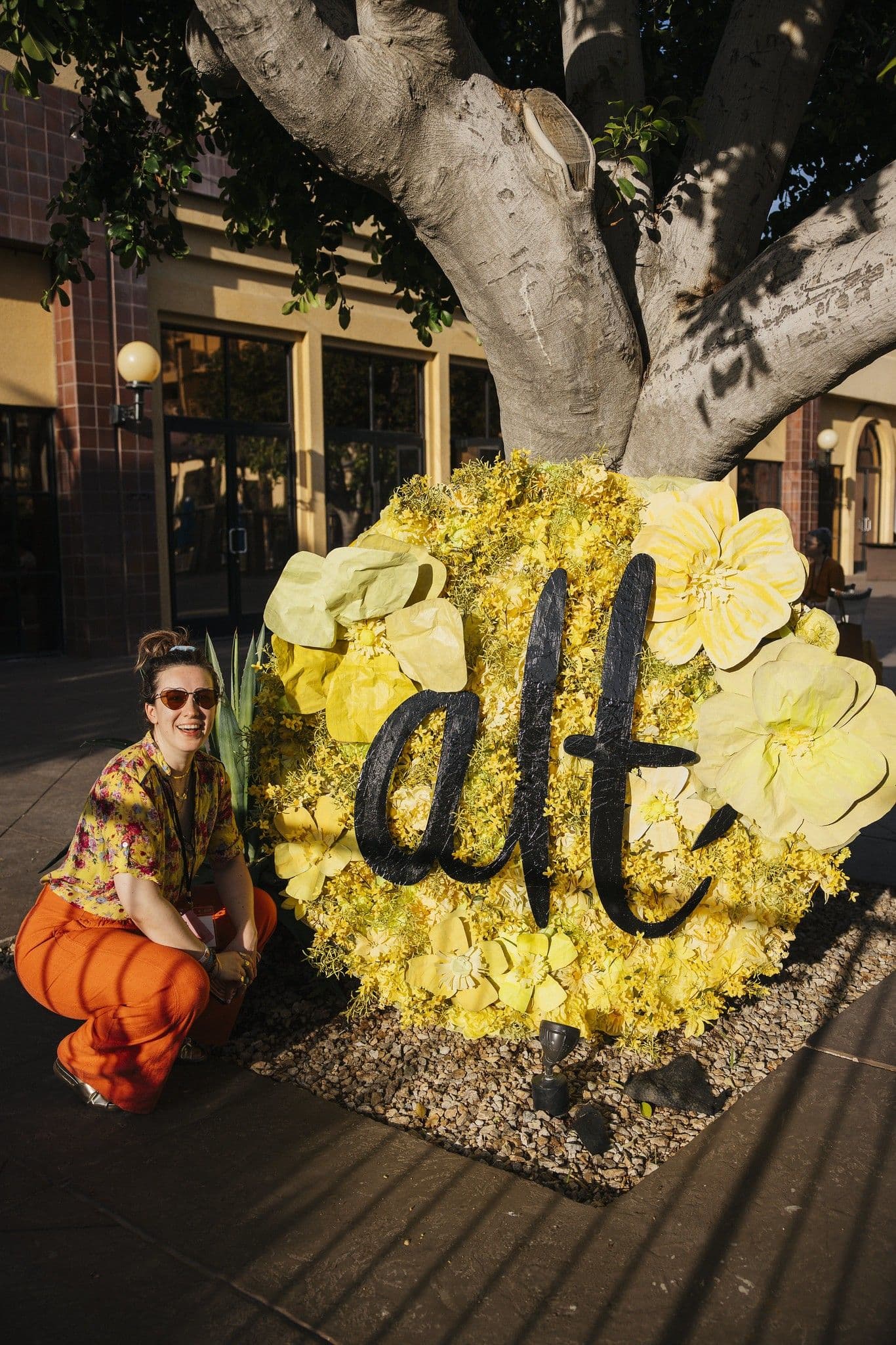 A person crouches beside a large circular floral display featuring bright yellow flowers and the word “alt” in bold black lettering, positioned at the base of a large tree in an outdoor plaza with warm sunlight.
