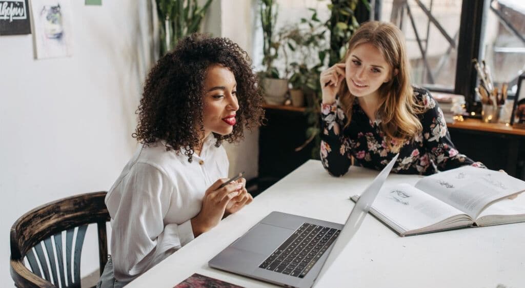 Two women sit a desk working on a laptop, planning a direct mail project.