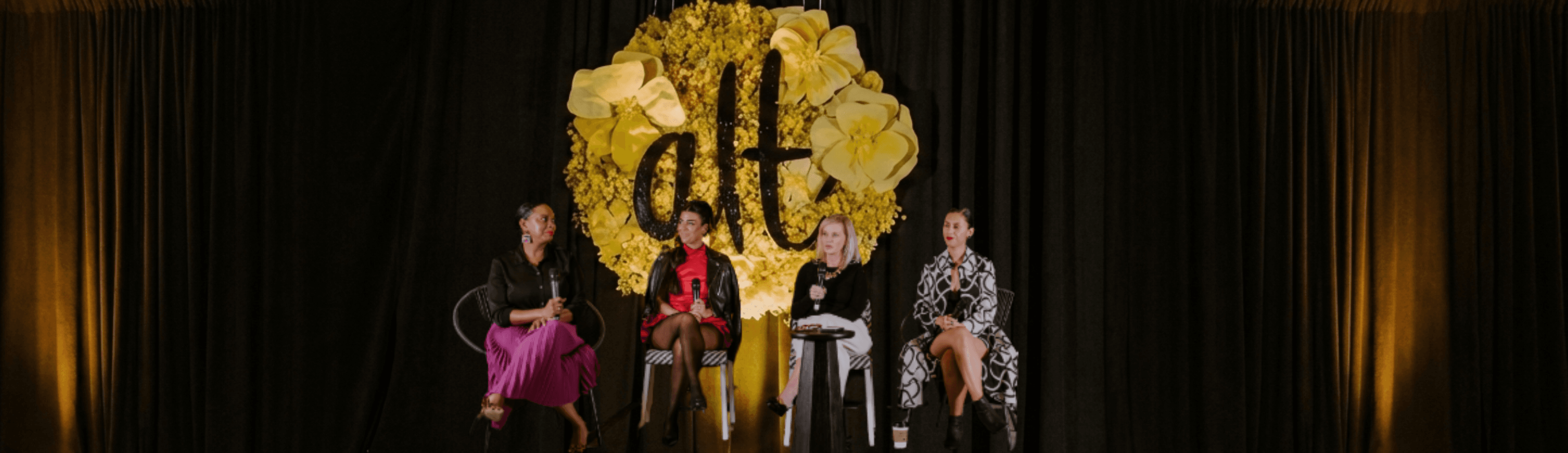 A panel of four speakers sits on a stage in front of a large floral backdrop featuring the letters “alt.” Each speaker holds a microphone, seated on modern black chairs, with a dark curtain behind them and warm event lighting highlighting the scene