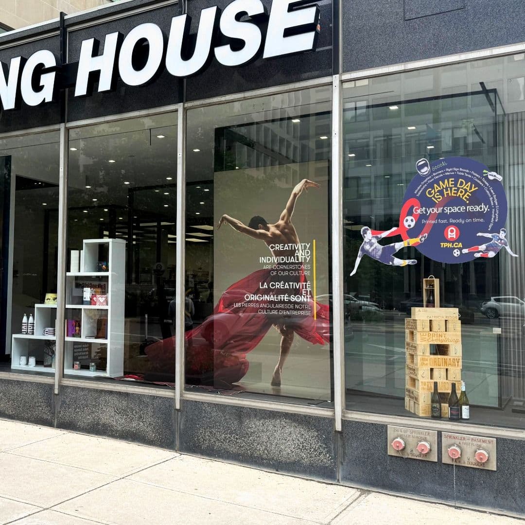 Storefront windows of a "ING HOUSE" building, featuring a dancer poster, white shelving with products, a "Game Day Is Here" soccer ad, and stacked wooden crates with bottles.