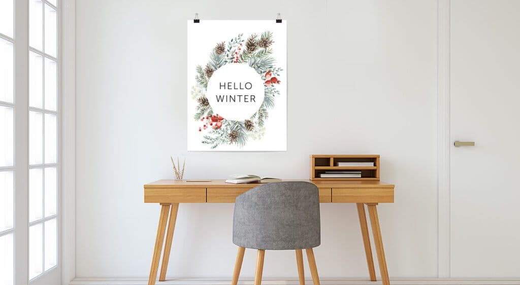 A large poster print hands above a minimalist home office desk. Poster features a graphic design of a winter wreath with the words "hello winter" typeset in the middle of the wreath.