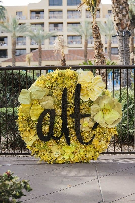 A large circular floral display made of bright yellow flowers and oversized blooms features the word “alt” in bold black lettering. It is positioned outdoors in front of a metal fence, with palm trees and a resort-style building in the background.
