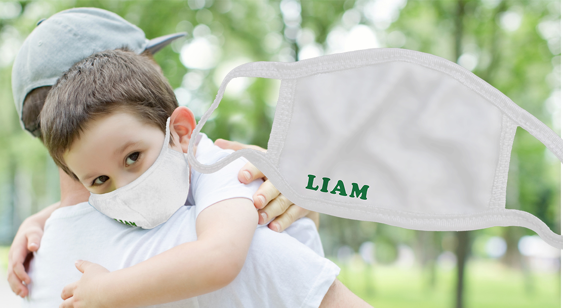 Photo of a young boy being embraced by his parent, wearing a fabric face mask with his name embroidered in it. Detail shot of the face mask with the embroidered name is next to it.