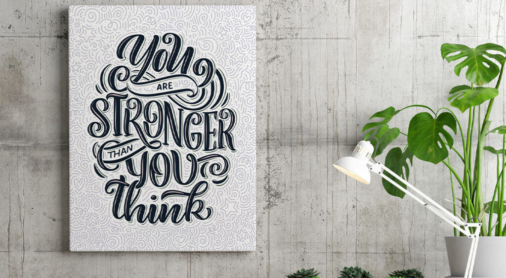 A canvas print features typographic art with the quote "You are stronger than you think" styled in black, grey and white above what appears to be a workstation with a lamp and a houseplant.