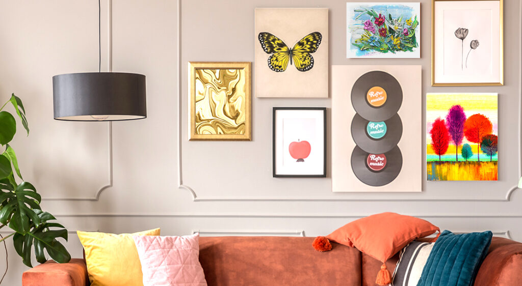 Cropped image of a living room wall, above a dark orange couch features an eclectic gallery wall of a butterfly canvas print, a collection of record albums printed on canvas, along with some framed prints of flowers, an apple, trees, and abstract swirls. the canvas and framed prints of different sizes are grouped closely together on the wall.