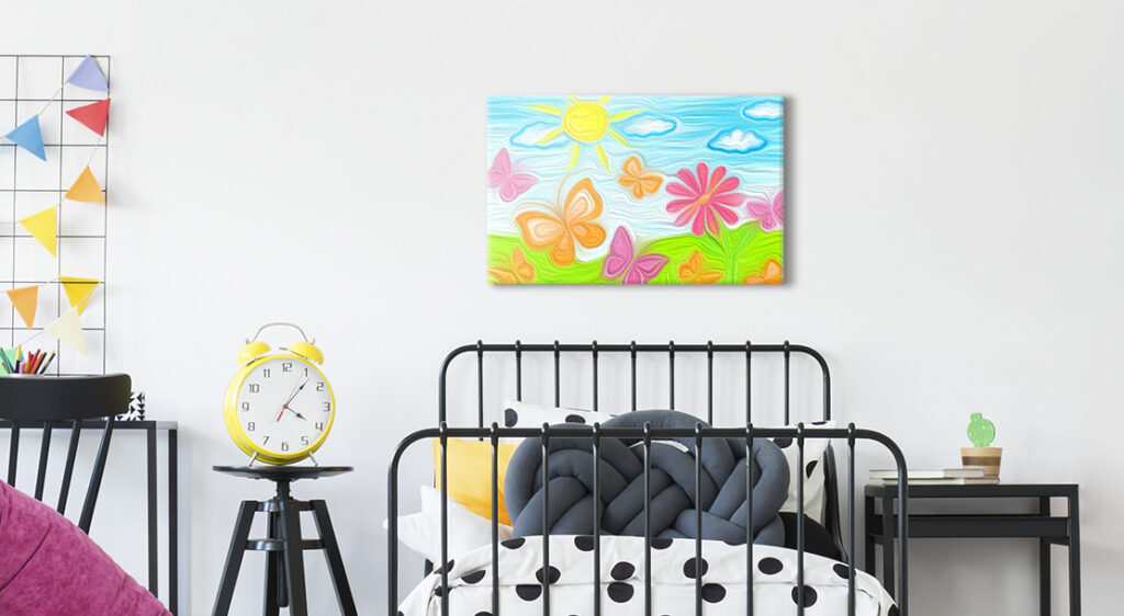 A medium sized canvas print of a child's drawing hangs above a small twin bed in a child's bedroom. The child's drawing is a bright landscape of green grass, blue sky and a bright yellow sun with large butterflies and a flower in the foreground. 