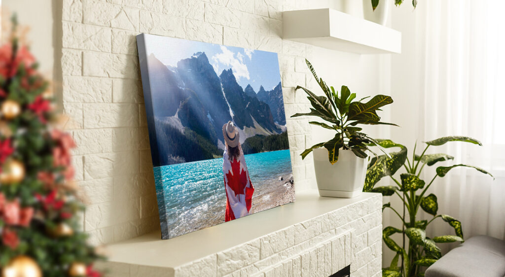 A canvas print photo leans on top of a white brick fireplace mantle beside large houseplants. The canvas is a photograph of an unknown glacier lake in Banff and centered in the image is a woman looking at the mountains beyond the lake with her back to the viewer, wrapped in a Canadian flag. The edge of a Christmas tree peeks out from the edge of the living room.