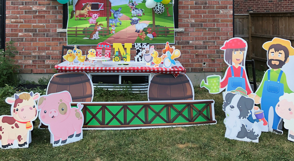 Photo of a children's party, with a farm theme. Cardboard cutouts of barnyard animals are in the foreground, and a table is made to resemble a barn. On the table is a cutout saying "ONE". Behind the table is another poster depicting a farm scene.
