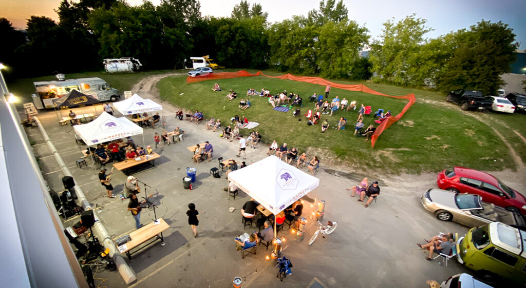 Birds eye view or drone image that shows the Market Brewing patio. Half parking lot and half grassy hill, customers enjoy the atmosphere under pop-up tents with picnic tables and lawn chairs on a summer evening.