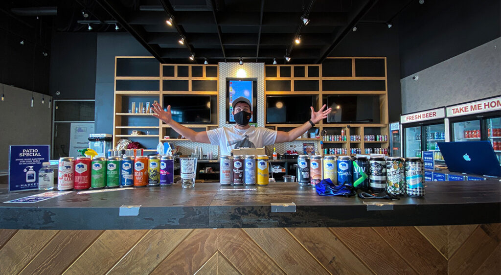 Masked bartender in baseball cap stands behind bar top with his arms spread wide showing off a display of the Market Brewing beer cans and bottle shop brews lined up side by side.