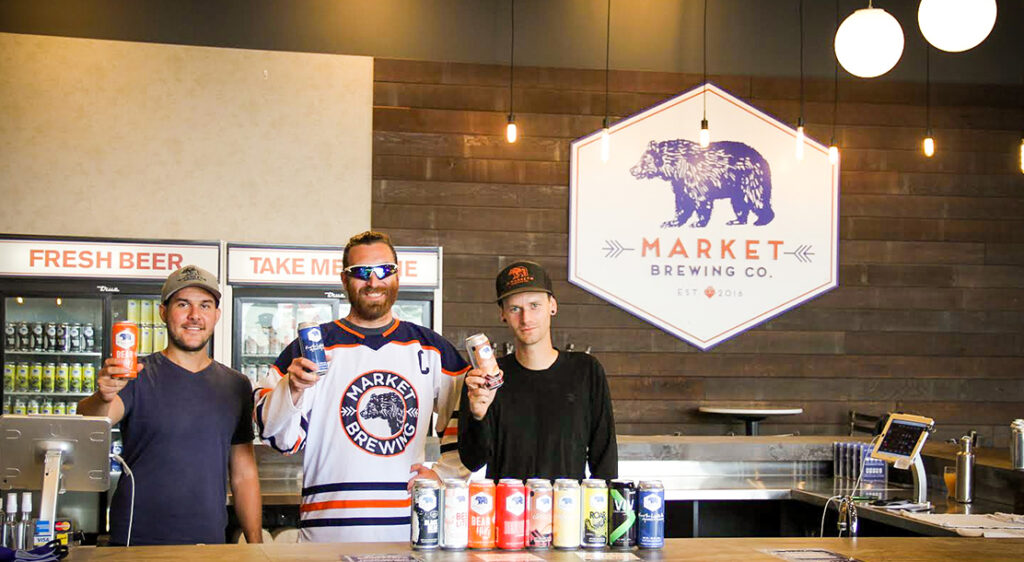 Three male staff members holding Market Brewing company beer cans stand together behind the bar top in the tap room. A large Market Brewing Company sign hangs in the background on a rustic wood wall.