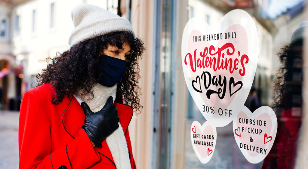 Bundled and masked woman on sidewalk of downtown district is looking at vinyl stick-on window decals in a retail storefront that advertise curbside pickup+ delivery, Valentine's Day sales, and gift card availability.